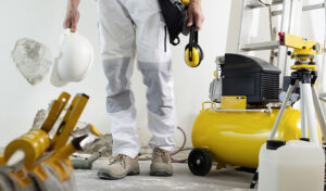 A Man In White Overalls Stands Next To A Yellow Air Compressor At A Restoration Site, Emphasizing Site Safety