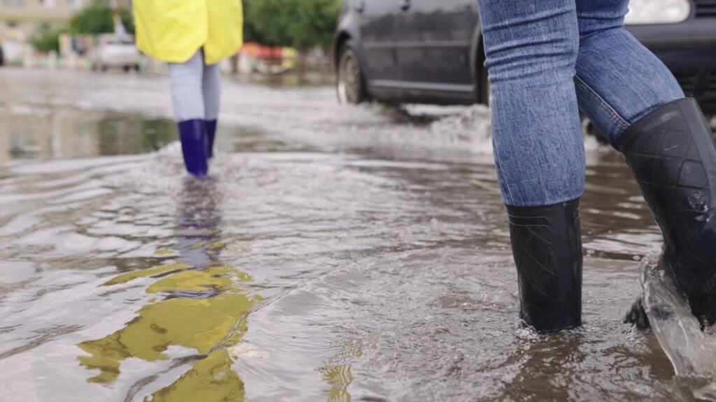 Two individuals in rain boots wading through a flooded street, braving the heavy rain.