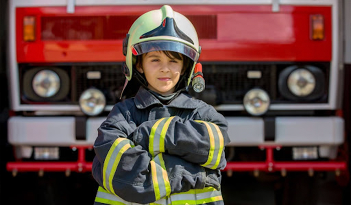 A young boy in a fireman's uniform standing in front of a fire truck, promoting fire safety.