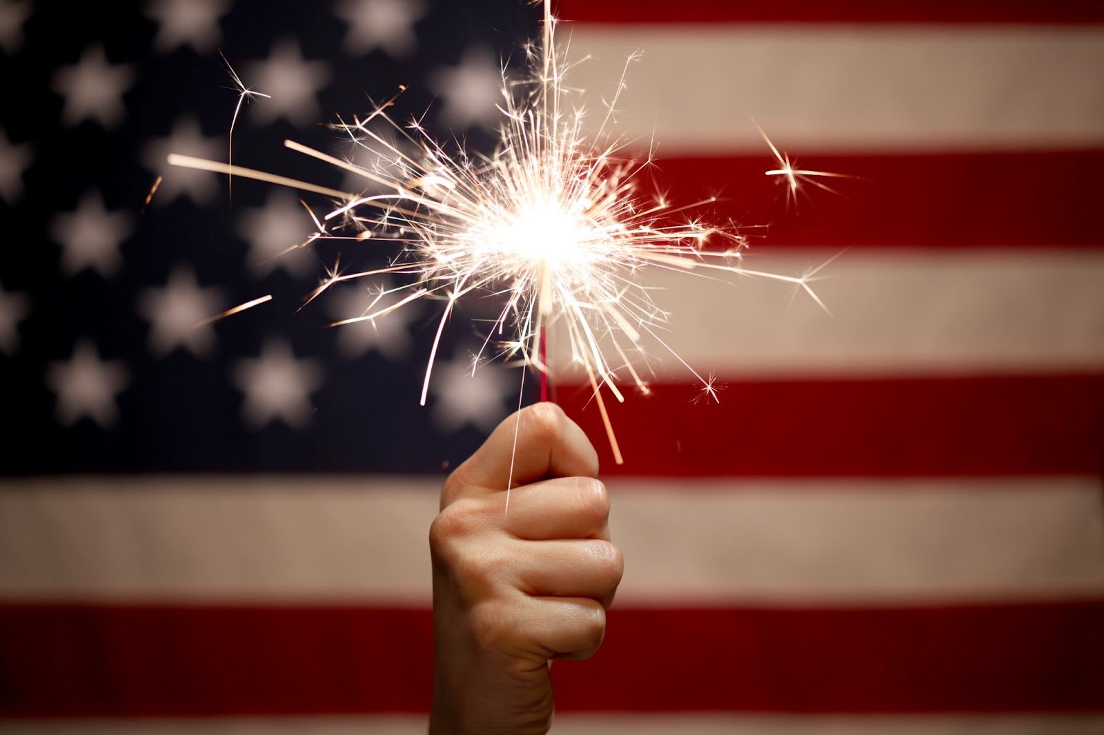 Hand holding sparkler in front of American flag, emphasizing fire safety.