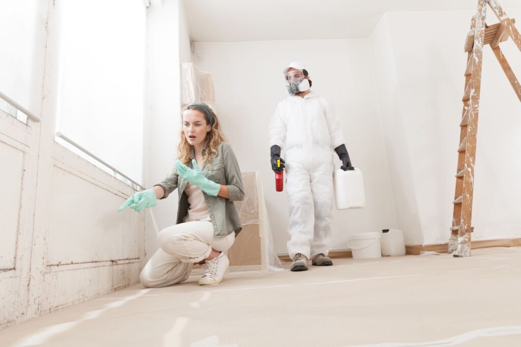 A man and woman in protective gear are painting a room, ensuring safety during mold remediation and removal efforts.